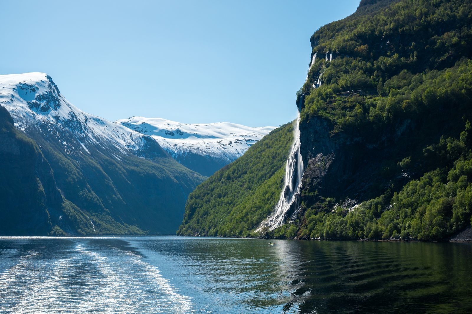 Waterfall pouring down a green cliff into a fjord, seen from a ship wake on a Norwegian fjords cruise